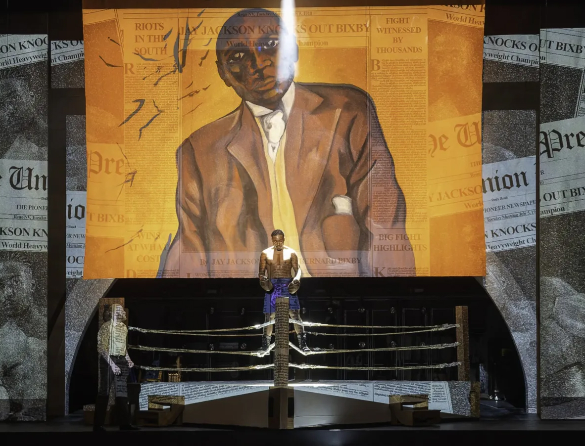 painting of a black man in a brown suit against a yellow background, hanging over a stage set with a boxing ring