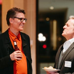 Two men laughing, holding drinks and hors d'oeuvres