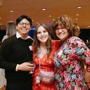 Man in black sweater and two women in brightly colored dresses smile for the camera