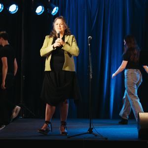 LTI Director Julie Gunn, wearing a black dress and lime jacket, speaks into a microphone on stage