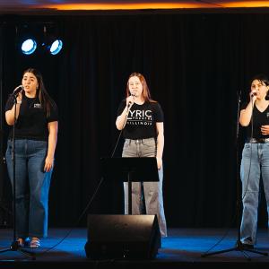 Three female lyric theatre students in black t-shirts sing into microphones on stage